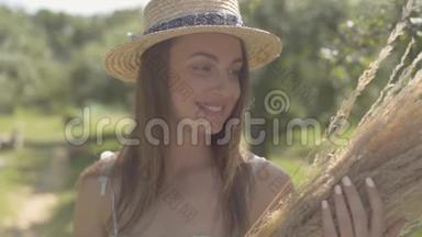 <strong>可爱</strong>的年轻女子戴着草帽，穿着长长的白色连衣裙，站在绿色的夏日花园里，手里拿着<strong>一堆</strong>干草。 农村